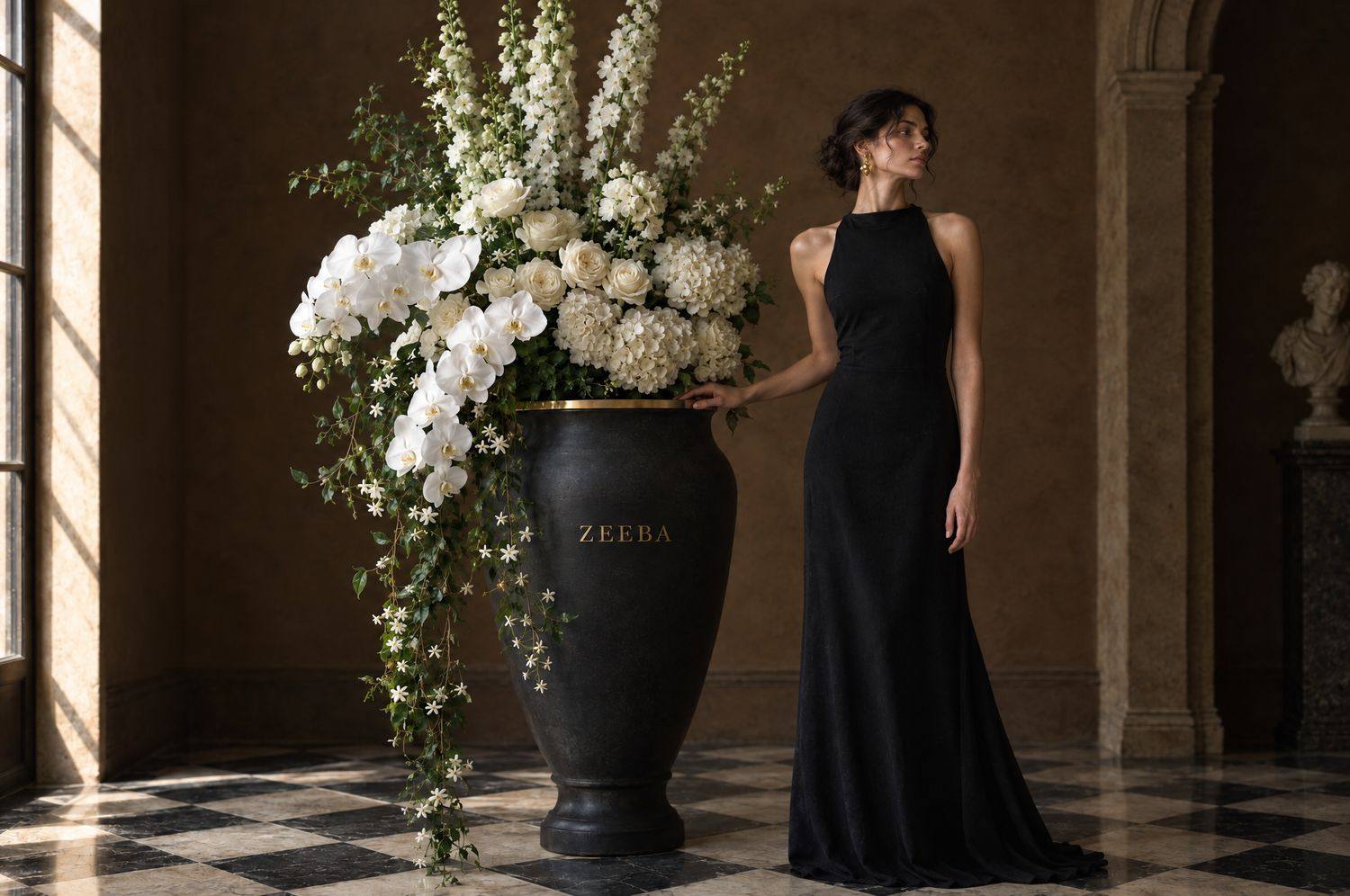 A woman in a black gown beside a tall black Zeeba urn of white orchids
