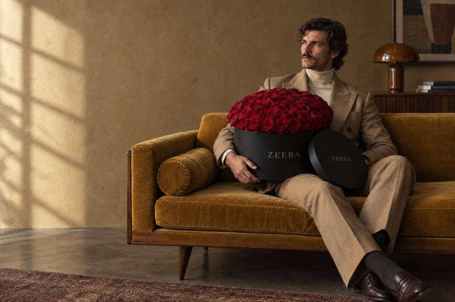 A man in a camel suit on a mustard sofa holding a black Zeeba hatbox of red roses